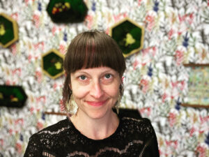 photo of a woman in her forties with brown chin-length hair and bangs, green eyes, dangle leaf earrings, and a black shirt with an out of focus background including moss art wall sculptures and a colorful leaf printed wallpaper.